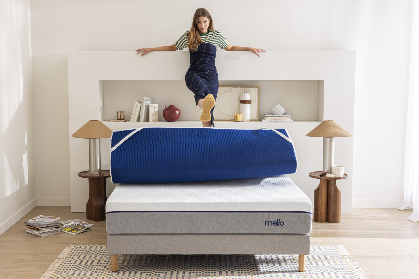 Une jeune femme souriante est debout sur son matelas et déroule son sur-matelas d'un coup de pied