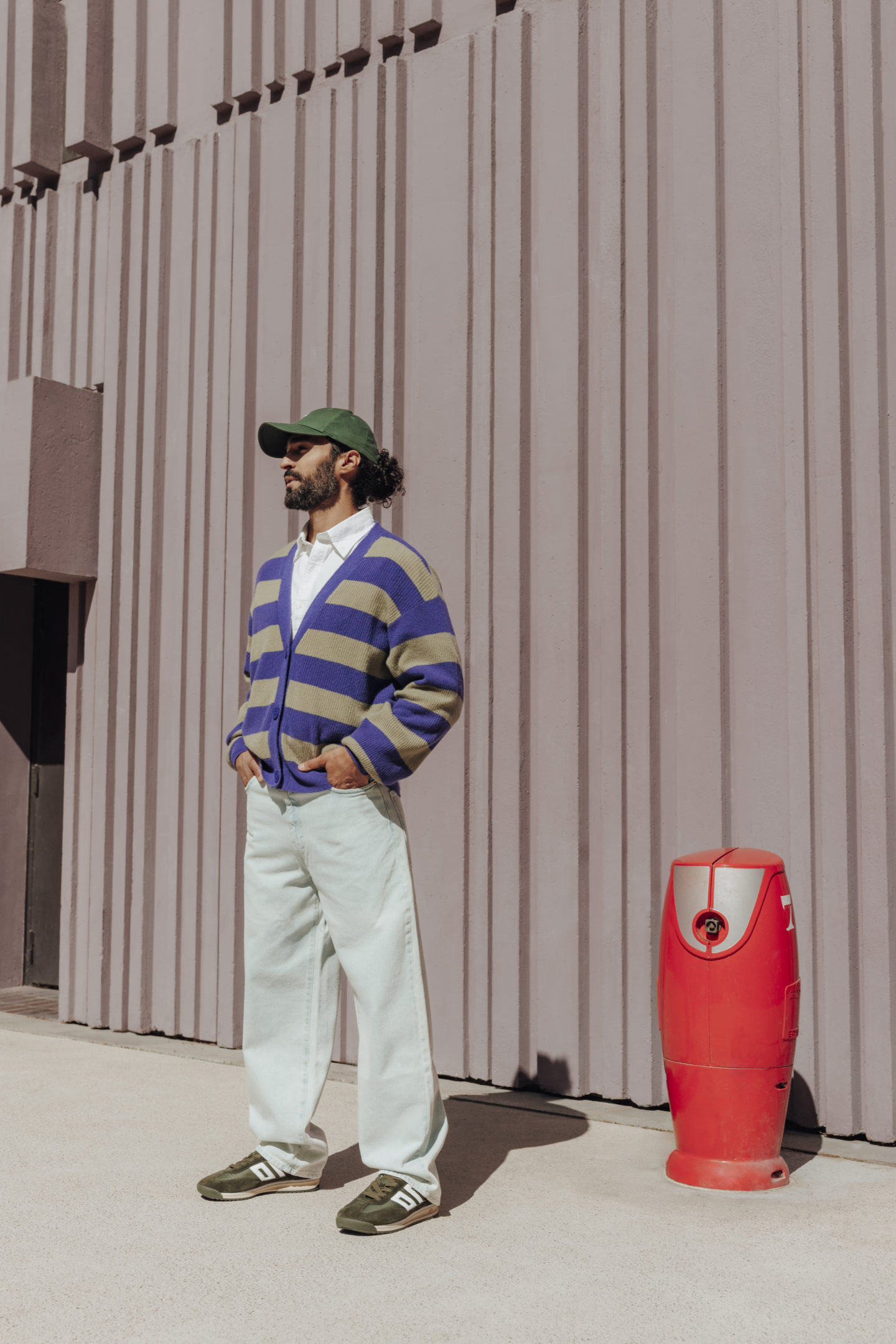 Homme debout de 3/4 avec une casquette verte, une tenue décontractée et des sneakers Jef devant un mur gros/mauve