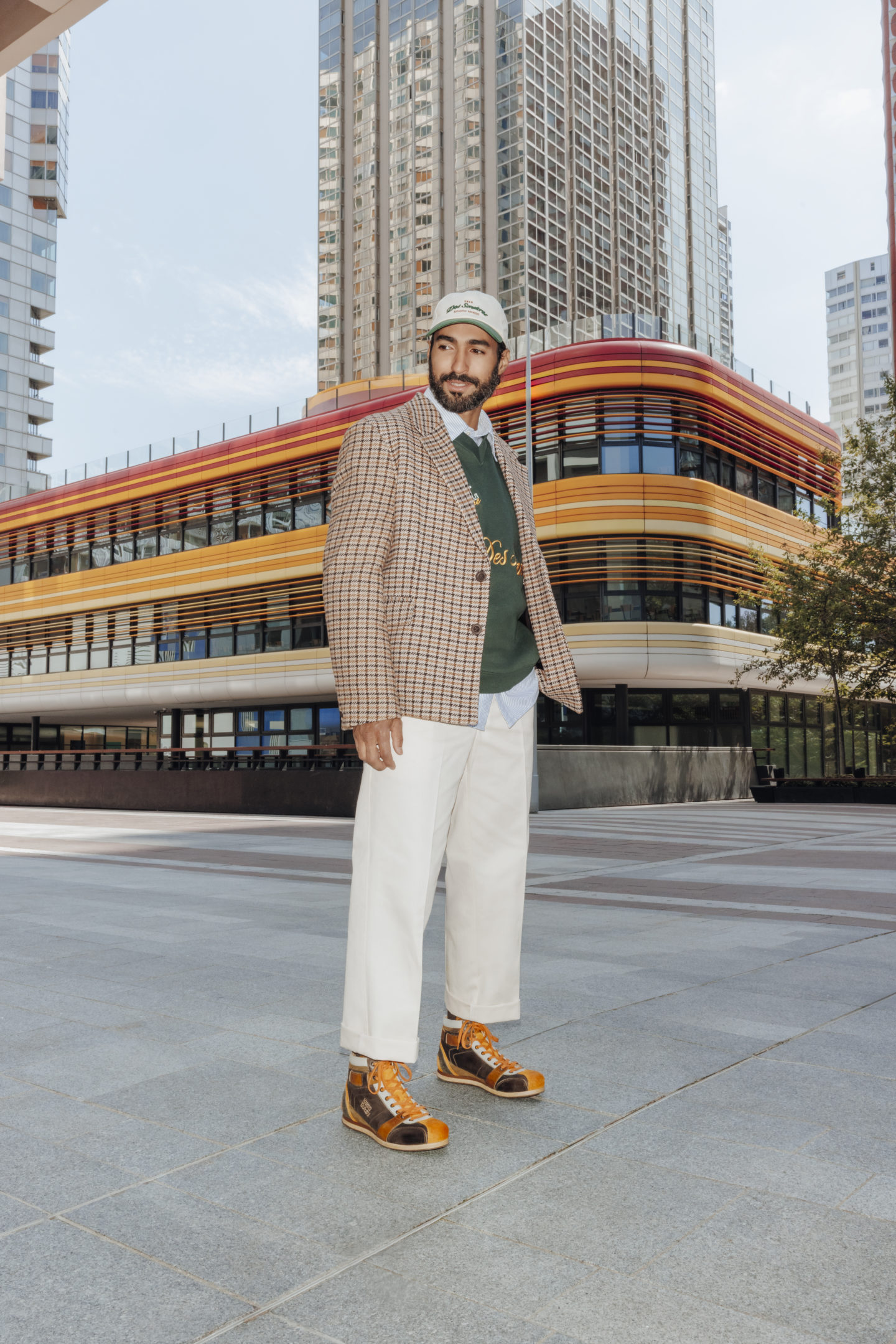 Homme qui se tient debout dans un environnement urbain, béton et bâtiment orange, portant une tenue dans les tons beiges et verts avec une paire de sneakers Jef