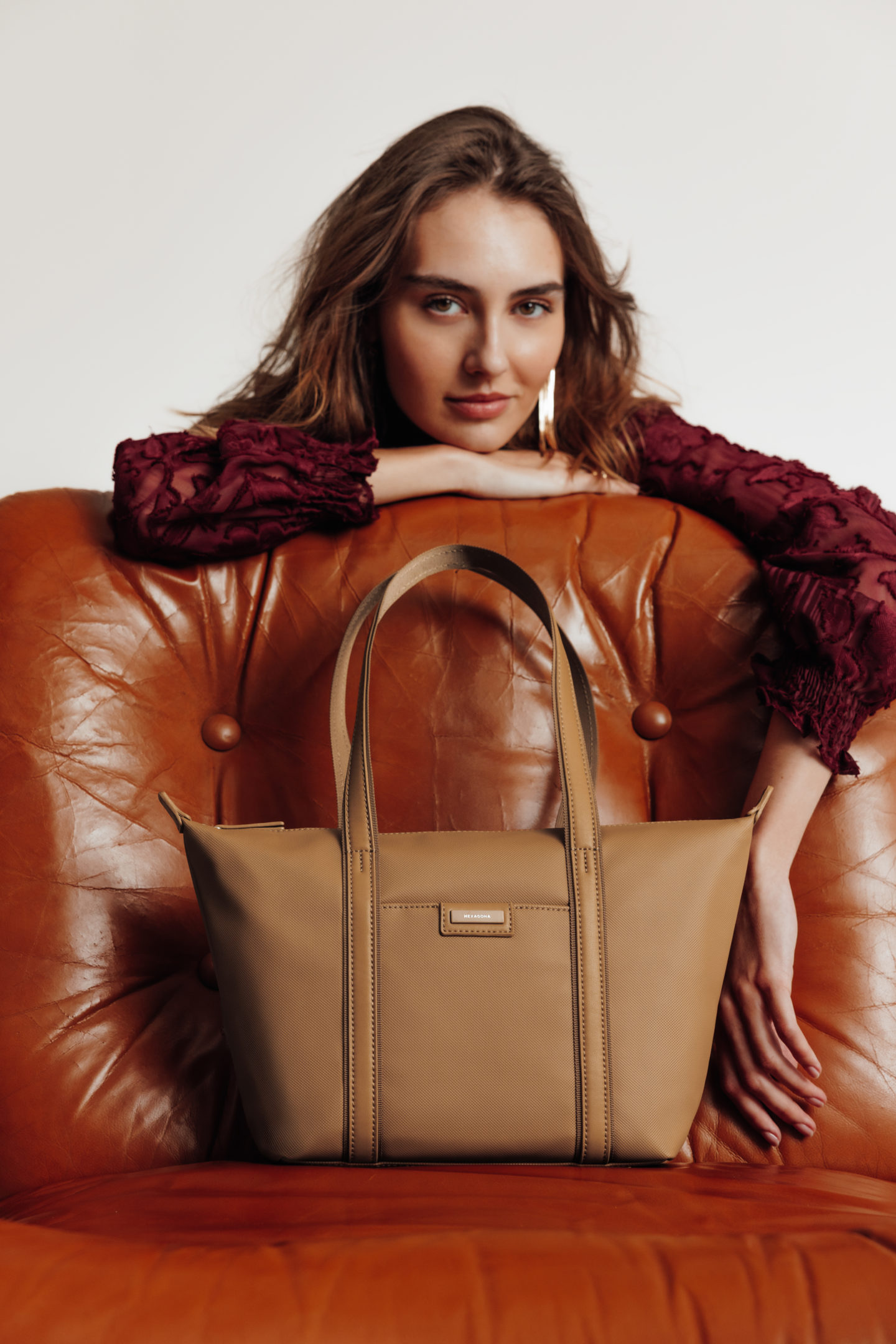 Zoom d'une jeune femme accoudée derrière un siège en cuir cognac avec un sac à main posé sur l'assise de ce siège avec un fond de studio blanc